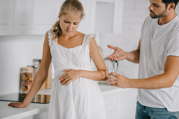 pregnant woman in white nightie having discomfort while husband giving medicines and glass of water to her at home