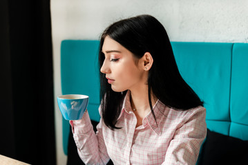 Beautiful brunette woman in pink checked shirt sitting in cafe with loft interior, resting and smiling. Pretty student female drinking coffee americano. Concept of freelancer, business.