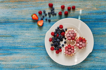 Cook cakes served on a white plate, tartlets with raspberry jam and fresh raspberries sprinkled icing sugar, strawberries on a plate next to raspberries and blueberries