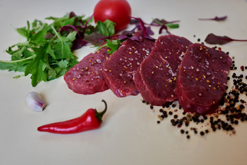 On a white table a smooth, fresh meat is cut into steaks beside mix of greens, garlic, tomatoes, red peppers from the buyout will be made salad