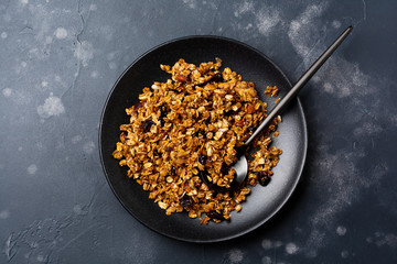 Oatmeal granola with dried fruit, chia seeds and sunflower, honey in black ceramic plate on dark background. Top view.