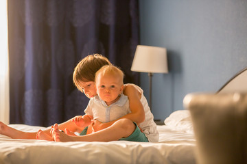 Two sibling children, baby boy and his older brother, sitting on bed in sunny bedroom, playing together