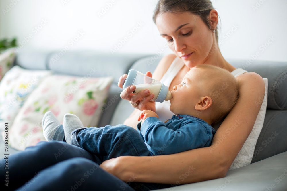 Poster Mother, feeding her baby boy from bottle, sitting on the couch at home