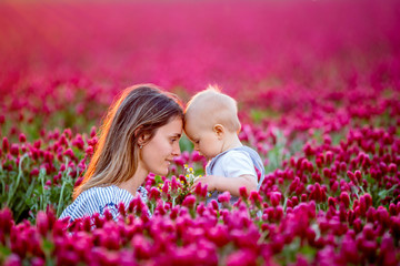 Young mother, embracing with tenderness and care her toddler baby boy in crimson clover field, smiling happily