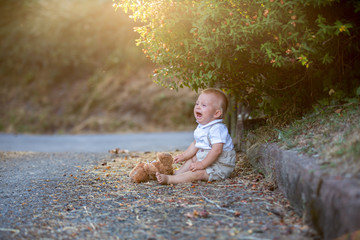 Sad little toddler boy, holding teddy bear, sitting lonely on a road in park, crying with sadness