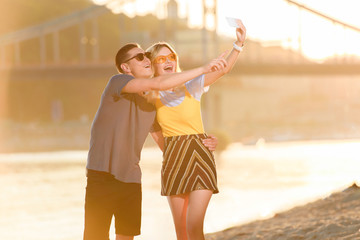 young couple taking selfie with smartphone on river beach during sunset