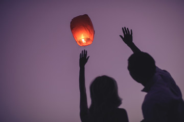 silhouettes of couple waving hands to flying chinese lantern in violet evening sky © LIGHTFIELD STUDIOS