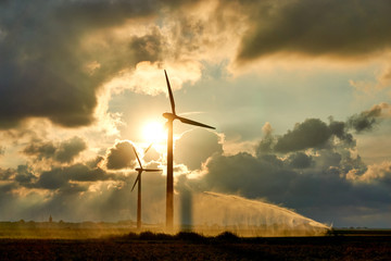 Two wind turbines and irrigating crop water gun or water spray at sunset in backlight against an...