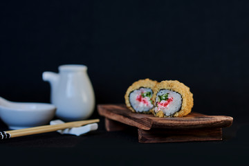 Traditional japanese food. Tempura sushi rolls made of smoked eel and crab meat, served with wasabi, ginger and soy sauce, on a brown wooden board for sushi on black background. Selective focus..
