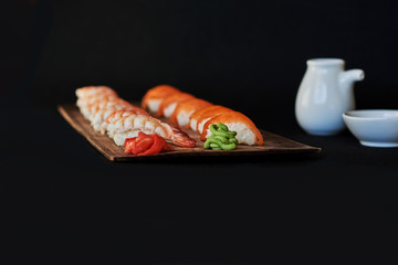 Sushi set with salmon, prawns, soy souce, wasabi and ginger on a brown wooden board for sushi on black background. Traditional Japanese cuisine. Delicious dinner in the restaurant. Selective focus.