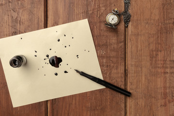 An overhead photo of an ink well with drops of ink and a nib pen, with copy space, on a dark rustic...
