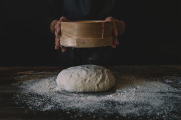 Woman sift the flour through a sieve to prepare the dough for pizza on a dark background. Sprinkling of dough with white wheat flour. Low key shot, close up on hands, some ingredients on table.