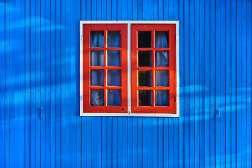 The old blue wall of a wooden house with a window is painted with red paint. Small red wooden window and ancient glass on the wall of old house, vintage building.