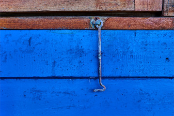 Old metal shutter hook on edge of  wooden window close-up. Hanging vertically in front of blue wall. The cutout of an old wooden shutter with a hook. Blue window metal latch detail with copy space.