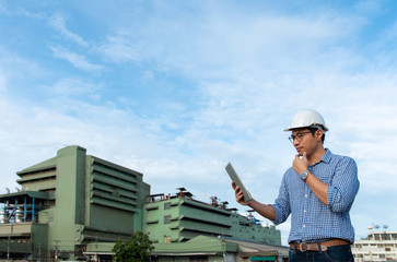 Engineer wear white hard hat looking tablet mobile in front of green factory, young male engineer using digital tablet outside industry