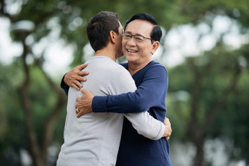 Aged Asian man smiling and embracing son while standing on blurred background of park