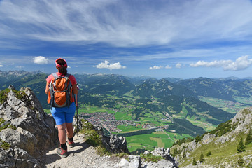 Rear view of a female hiker close to the Rubihorn mountain above the village Oberstdorf. Allgau Alps, Bavaria, Germany. Alpine landscape with rocky mountains, forests and blue sky.
