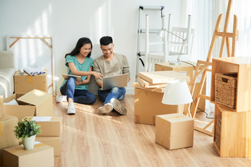 Cheerful Asian couple sitting on floor among carton boxes and browsing digital devices while relocating into new apartment together