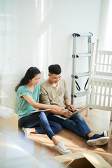 Young Asian man and woman smiling and browsing laptop while sitting on floor in new apartment