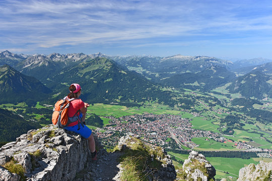 Female hiker at the summit of Rubihorn mountain enjoying the view to the village Oberstdorf and the Allgau Alps. Bavaria, Germany. Alpine landscape with rocky mountains, forests and blue sky.