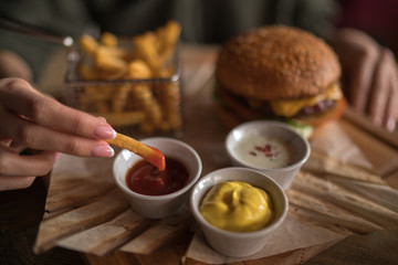 Woman holding cheeseburger and refreshing drink. Young girl holding in hands fast food burger, american meal. Beef burger in hands with french fries on rustic wooden background. Selective focus.