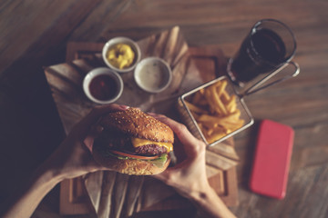 Woman holds burger with hands and fries on the background in cafe. Fresh burger cooked at barbecue in craft paper. American food. Big hamburger with meat and vegetables closeup unfocused at background