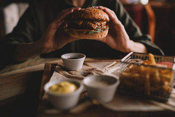 Woman holds burger with hands and fries on the background in cafe. Fresh burger cooked at barbecue in craft paper. American food. Big hamburger with meat and vegetables closeup unfocused at background