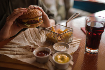 Woman holds burger with hands and fries on the background in cafe. Fresh burger cooked at barbecue in craft paper. American food. Big hamburger with meat and vegetables closeup unfocused at background