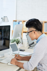 Side view of Asian man sitting at desk near computer and drawing in notebook while working in modern office