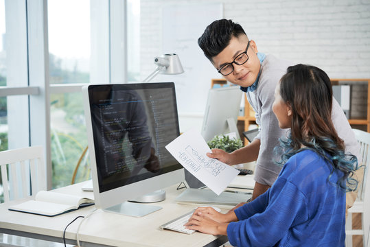 Side view of two Asian designers using computer and looking at each other while working on project together in stylish office