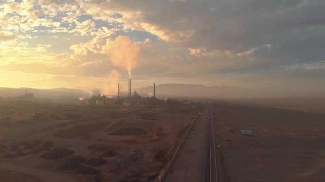 Aerial desert sunset with copper mine factory with smoke, pollution and colorful sky. Atacama Chile. Drone rising up filming during beautiful evening. Empty vast landscape with warm color grade.