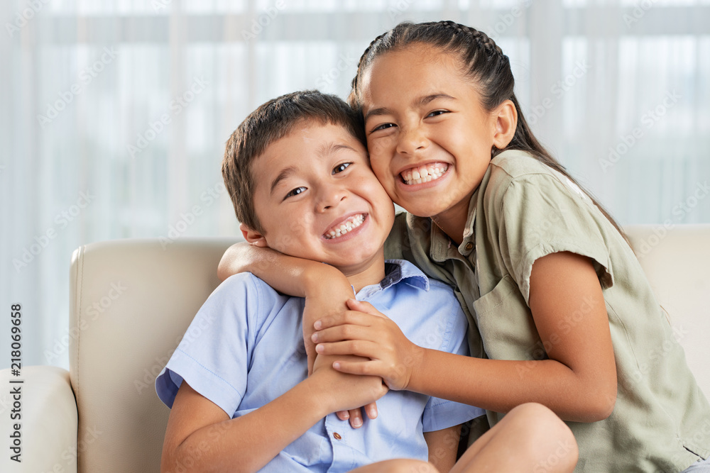 Wall mural Cheerful Asian girl smiling and embracing cute boy while sitting on comfortable couch together