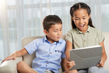 Asian boy and girl sitting on comfortable sofa together and looking at modern tablet with excited face expression