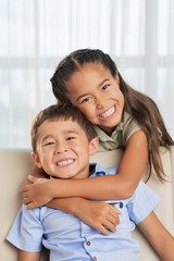 Sweet Asian girl smiling and looking at camera while hugging cute brother from back