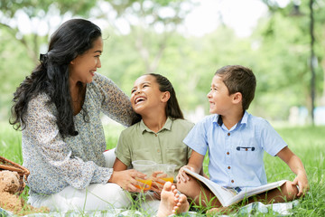 Beautiful Asian woman and two cheerful kids drinking and reading book during picnic in park on sunny day