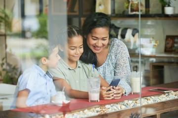 Fototapeta premium Asian woman and children smiling and browsing smartphone while sitting behind window glass in cafe