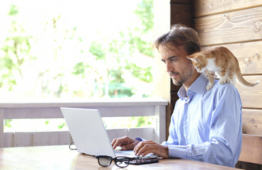 Young businessman is working on a laptop on an open terrace, kitten sits on his shoulder. Freelance concept © Gargonia