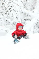 Fototapeta na wymiar Cute toddler boy in winter suit sitting on a big snowdrift, surrounded by snow-covered trees. Snowy winter. Copy space