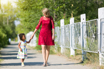 Naklejka premium A white woman is walking along the street with a black girl. A European mother with an African daughter in summer dresses is walking with her hand.