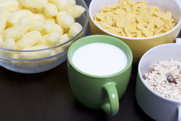 Useful breakfast. Muesli, corn sticks and flakes in cups. There's a cup of milk. On a dark background.