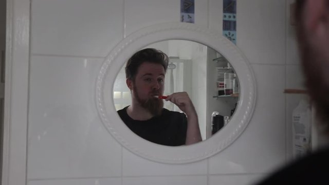 Tired white man with beard brushing teeth in morning viewed from reflection of bathroom mirror. Man is half asleep and looking tired.