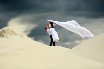 Mężczyzna podnosi wysoko dziewczynę w białej sukni z welonem na piaszczystej wydmie. - obrazy, fototapety, plakaty