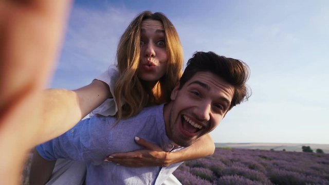 Young handsomw brunette man giving piggyback ride to hus beautiful lady. Lovers making selfie with camera in lavender field