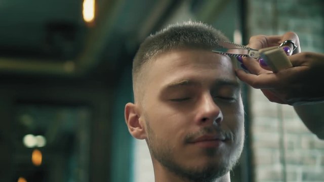 Professional hairdresser is cutting male's hair with scissors at barber shop. Smiling youg man is sitting on chair in hair salon and getting his hair cutted. Slow motion