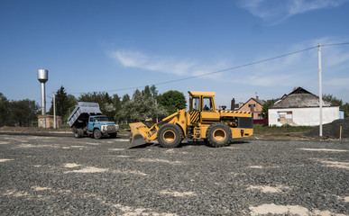 Yellow loader with empty bucket and blue truck standing on a stones at road construction and repairing asphalt pavement works. The stones for the road. Repaving.