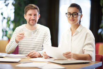 Adult woman and man with coffee cup sitting at table with papers and smiling at camera in modern office 
