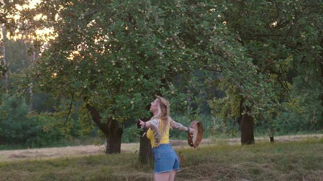 young blond hippie woman dancing with a tambourine on the grass in the park at sunset. Warm summer day and soft backlight. happy woman is dancing. the middle plan. a hot summer day. Slow motion