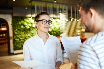 Adult woman and man holding papers and talking in leisure standing in light modern interior