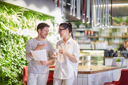 Stylish woman with coffee cup and man with papers standing in cafeteria and chatting in leisure