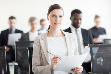 Adult elegant woman holding pencil and paper while sitting in conference hall with coworkers and...